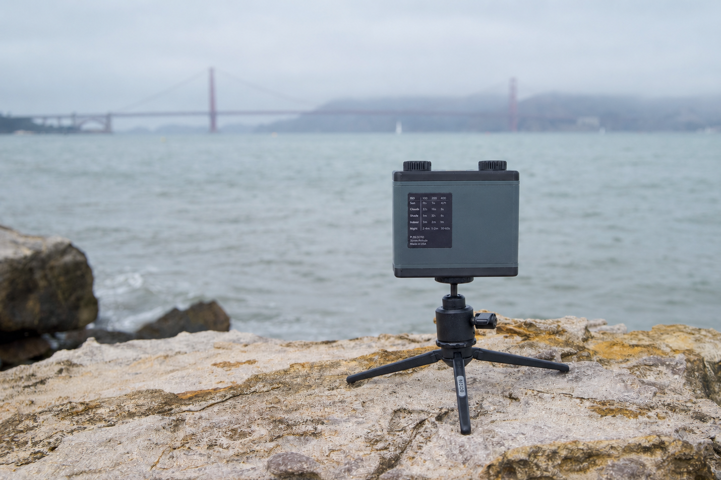 A black pinhole camera mounted on a small tripod sits on a rocky shoreline with water and a distant bridge in the background.